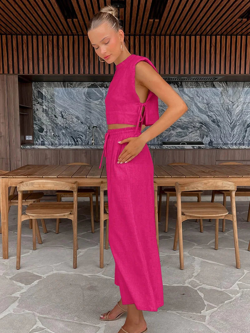 Woman in a bright pink outfit standing in an outdoor setting with wooden tables and chairs.
