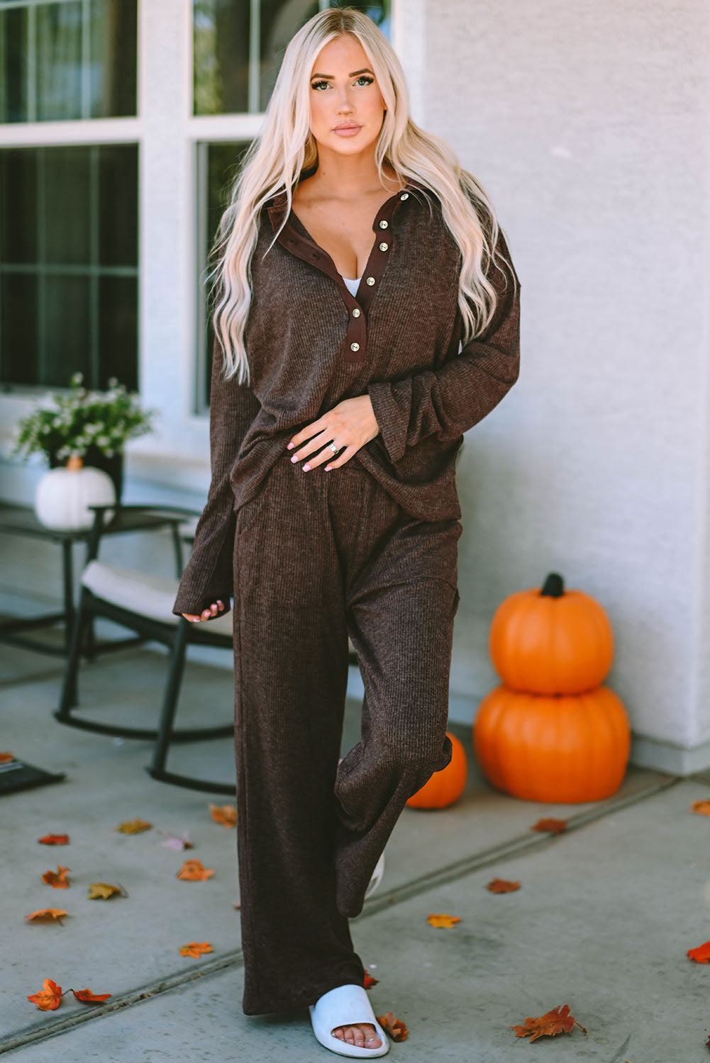 Woman in a brown outfit standing on a porch with pumpkins and leaves.