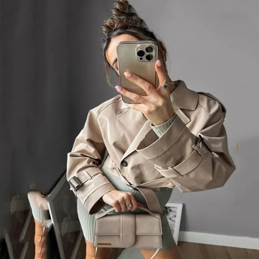 Person wearing a beige trench coat and holding a phone, standing in a room with a gray wall.