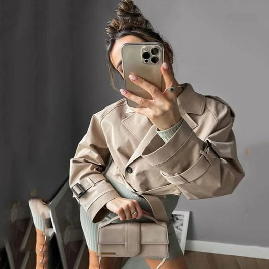 Person wearing a beige trench coat and holding a phone, standing in a room with a gray wall.